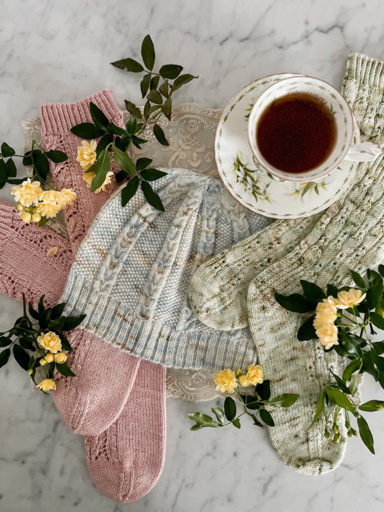 A top-down image of a pair of pink socks, a light blue hat, and a pair of green socks all laid flat on a white marble countertop. They're surrounded by tiny yellow roses and a white teacup full of tea.