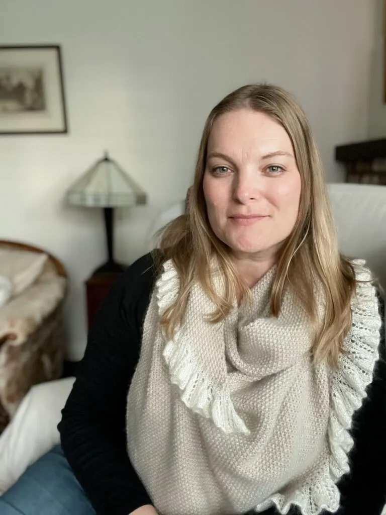 A blonde woman in her late 30s (that's me) wears a gray, seed stitch shawl with a white mohair ruffle. She's smiling softly at the camera.