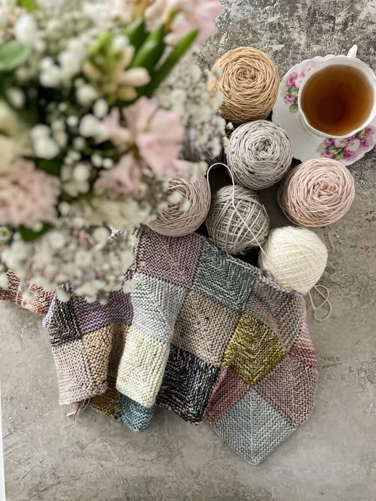 A top-down photo looking at a mitered square blanket in lots of pastel colors, a few balls of pastel and neutral yarn, and a pink and white teacup full of tea. Blurred in the foreground is the top of a vase full of pink and white hyacinths and baby's breath.