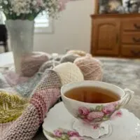 An ornate, antique teacup with pink flowers painted on it sits on a matching coaster. It's full of one of my favorite teas, Harney & Sons' Vanilla Comoro. In the background is a mitered square blanket made of lots of pastel yarns and a vase full of pink and white hyacinths and baby's breath.