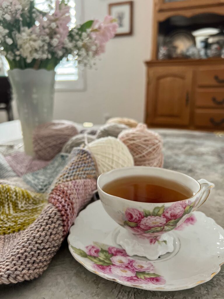 An ornate, antique teacup with pink flowers painted on it sits on a matching coaster. It's full of one of my favorite teas, Harney & Sons' Vanilla Comoro, which is one of this month's sip and stitch recommendations. In the background is a mitered square blanket made of lots of pastel yarns and a vase full of pink and white hyacinths and baby's breath.