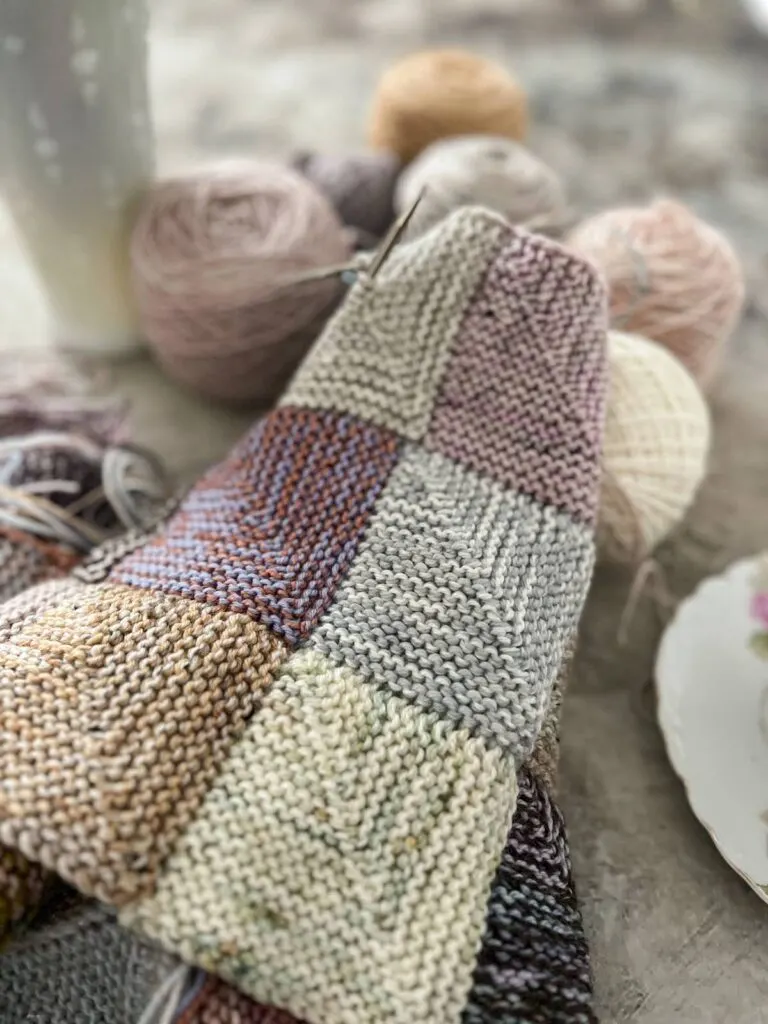 A close-up on several squares of my pastel mitered-square blanket. There are squares in pale green, ice blue, berry mixed with a soft pink, a wheat color held double with a tweedy gray, and more. This blanket is how I'm using up my many, many fingering-weight leftovers.