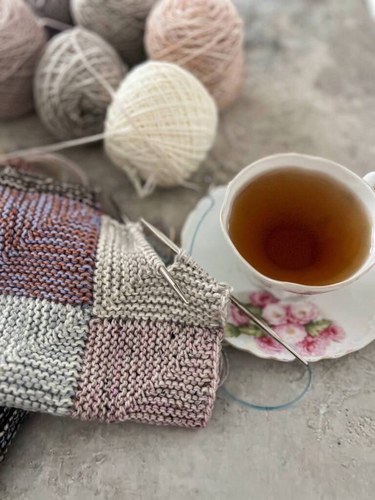 A close-up of an ornate, antique teacup with pink flowers painted on it and on the matching saucer. It's full of Harney and Sons' Vanilla Comoro tea. To the left of the teacup is my pastel mitered-square blanket, with a square in progress on a pair of steel needles (Addi Turbos, my forever favorites).