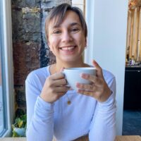 A young woman with light brown hair grins while holding a coffee cup near a window. Teresa has soft bangs that fall across her forehead and a happy, relaxed demeanor.