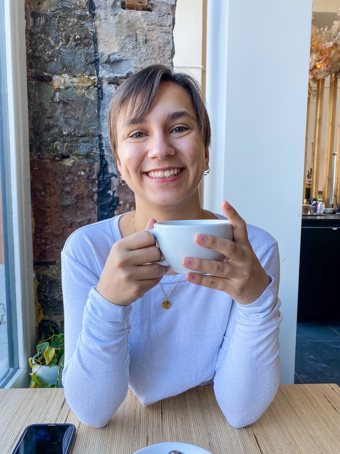 A young woman with light brown hair grins while holding a coffee cup near a window. Teresa has soft bangs that fall across her forehead and a happy, relaxed demeanor.