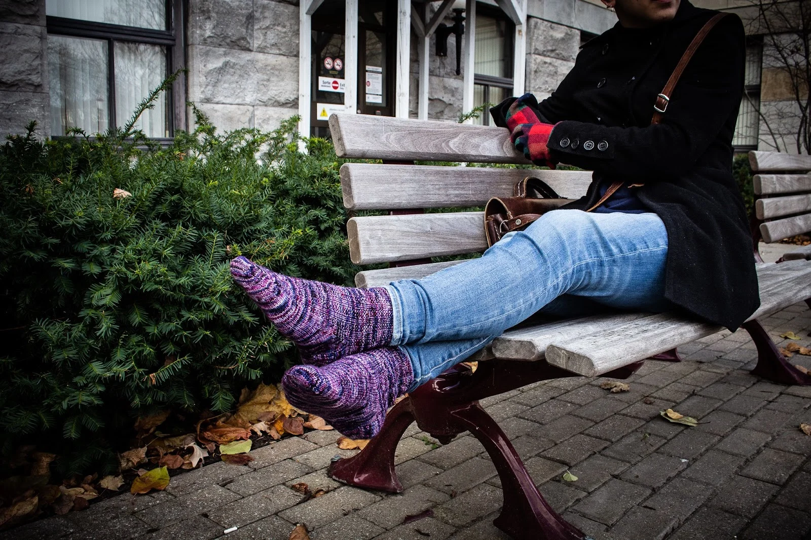 A pair of purple handknit socks on two feet that are crossed at the ankles. The model is sitting on a wooden park bench outside.