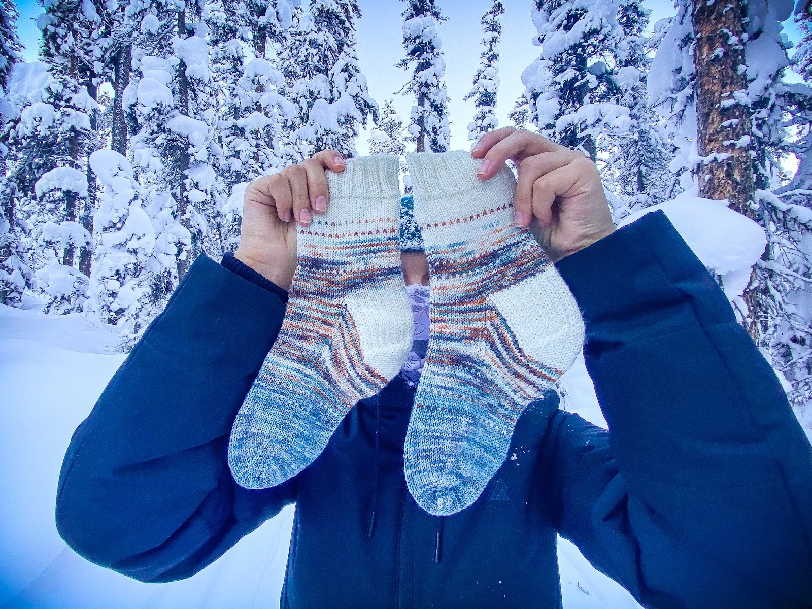 A person holds up two striped handknit socks in the middle of a snowy forest. The socks are knit with a gently variegated white, blue, and orange yarn, and the toes are pointing to the left.