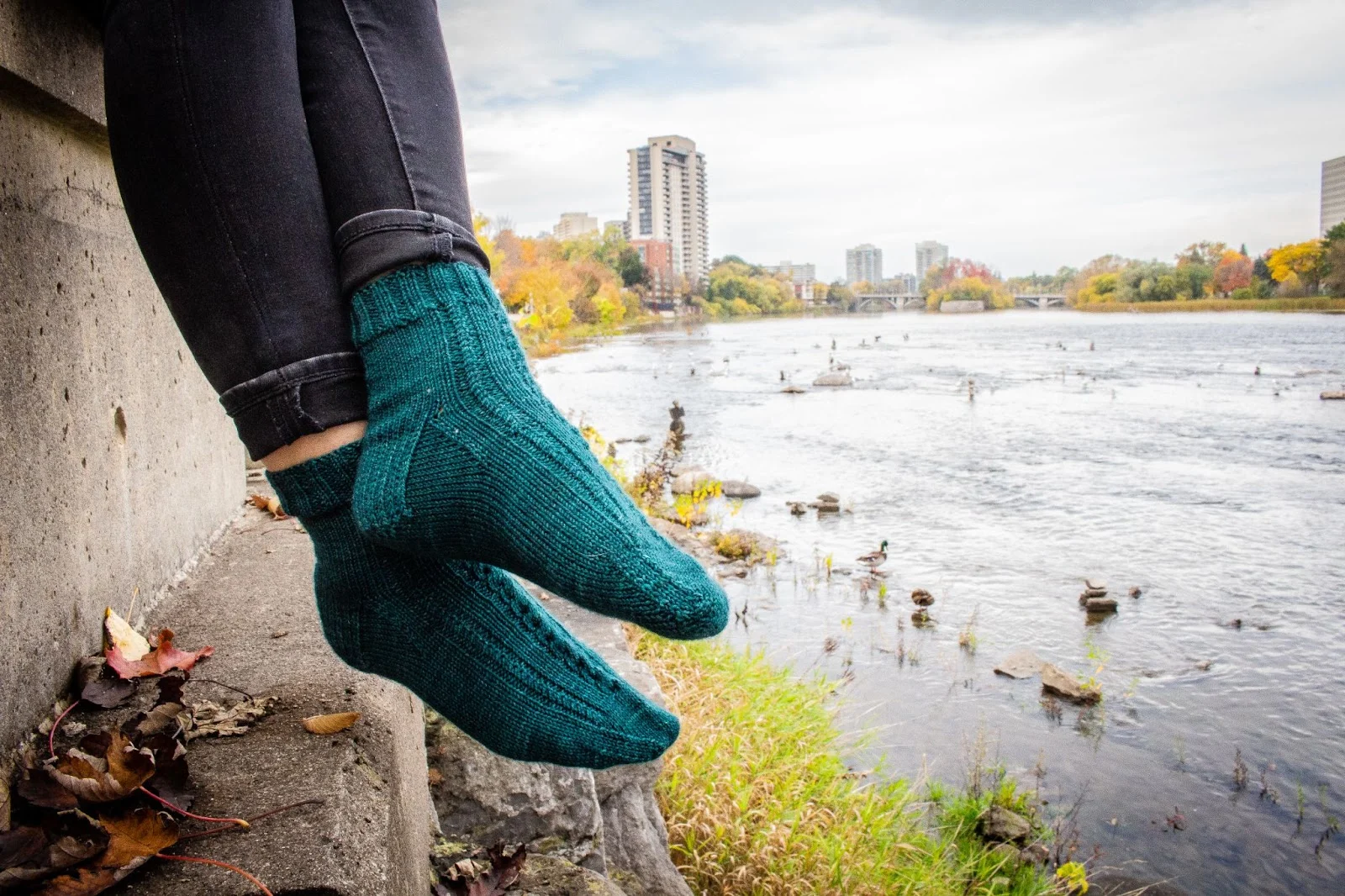 A pair of turquoise handknit socks with delicate cabled details are modeled by a woman sitting next to a river. Her legs are crossed at the ankles and the photo ends just below her knees.