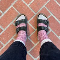A top-down photo of a woman's feet wearing purple lacy knit socks with Birkenstock sandals and black cropped jeans.