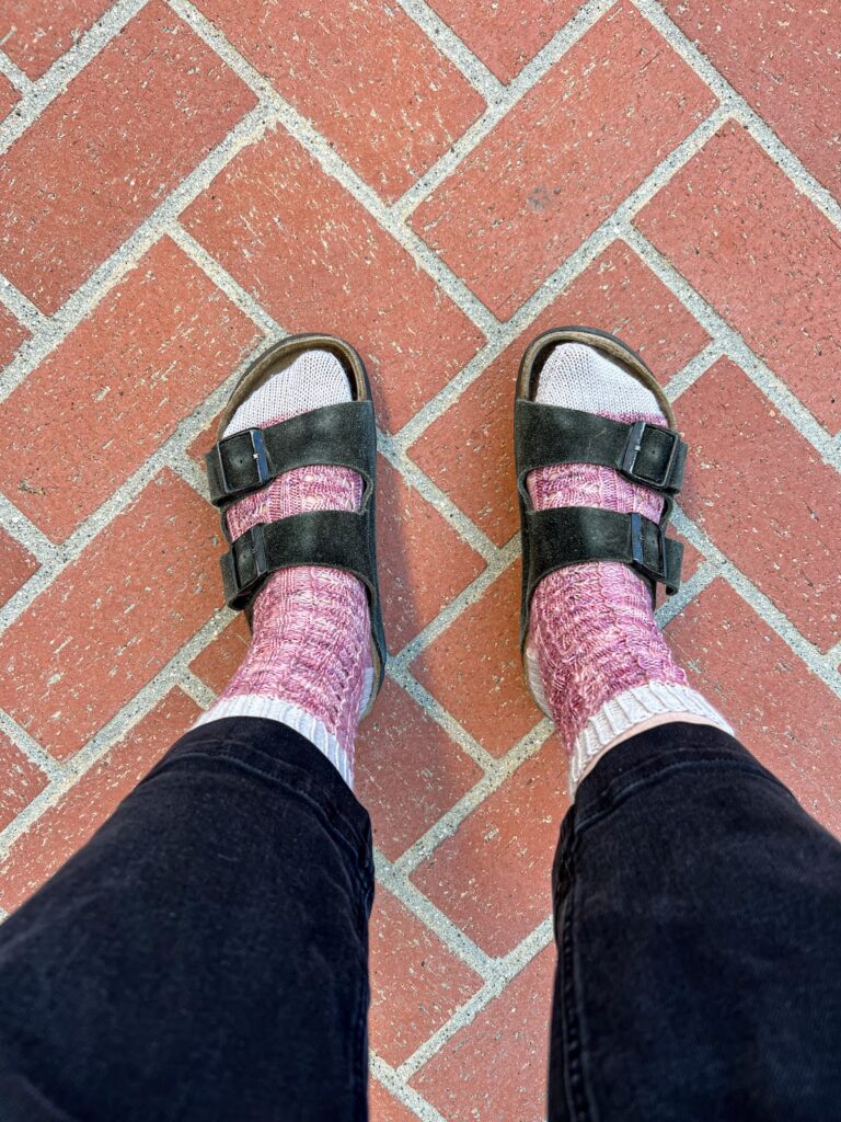 A top-down photo of a woman's feet wearing purple lacy knit socks with Birkenstock sandals and black cropped jeans.