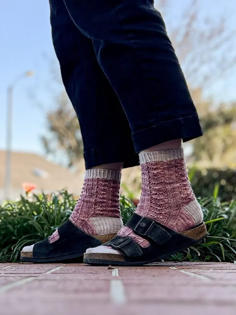 A pair of pinky purple lace socks with gray cuffs, heels and toes are worn by a woman with Birkenstock sandals over them. The photo focuses on her feet, which are mid-step. The photo is taken outside on a sunny day.