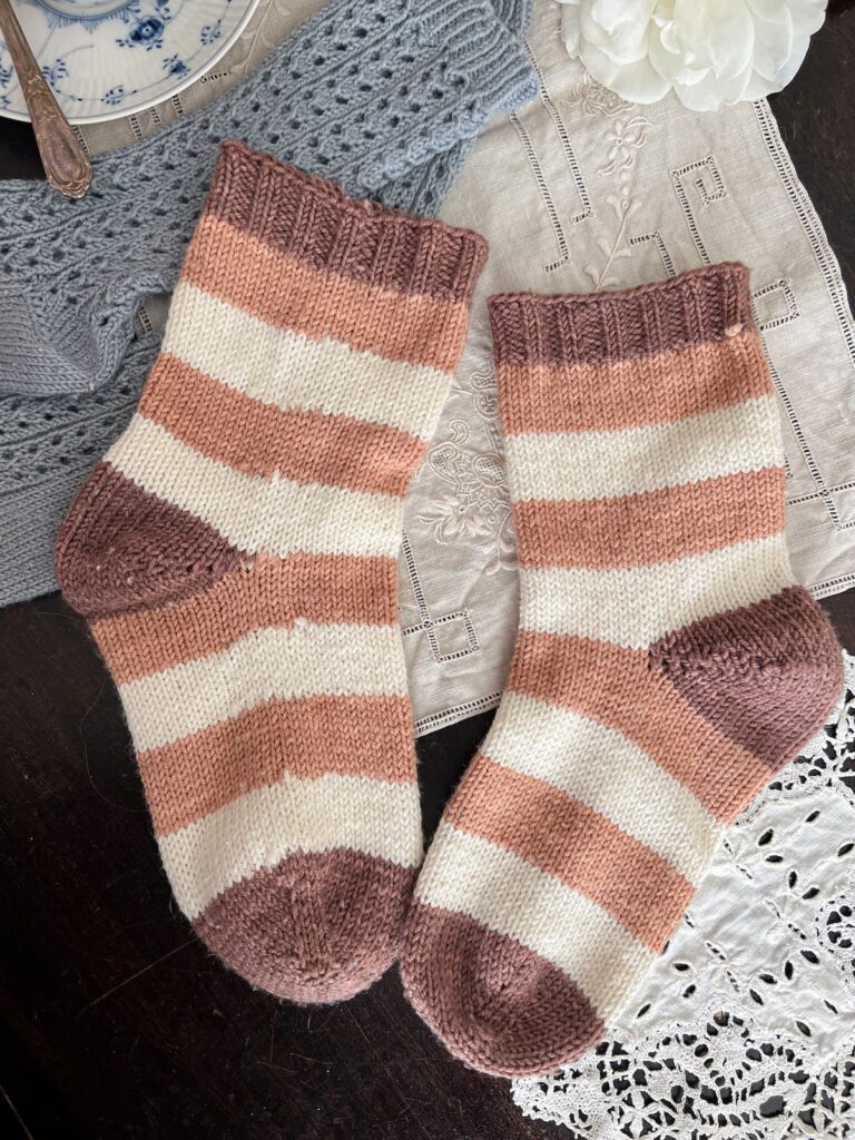 A pair of brown, pink, and white striped socks are laid out flat with the toes pointed inward. The sock on the right has had the pills removed, while the sock on the left still has significant pilling.