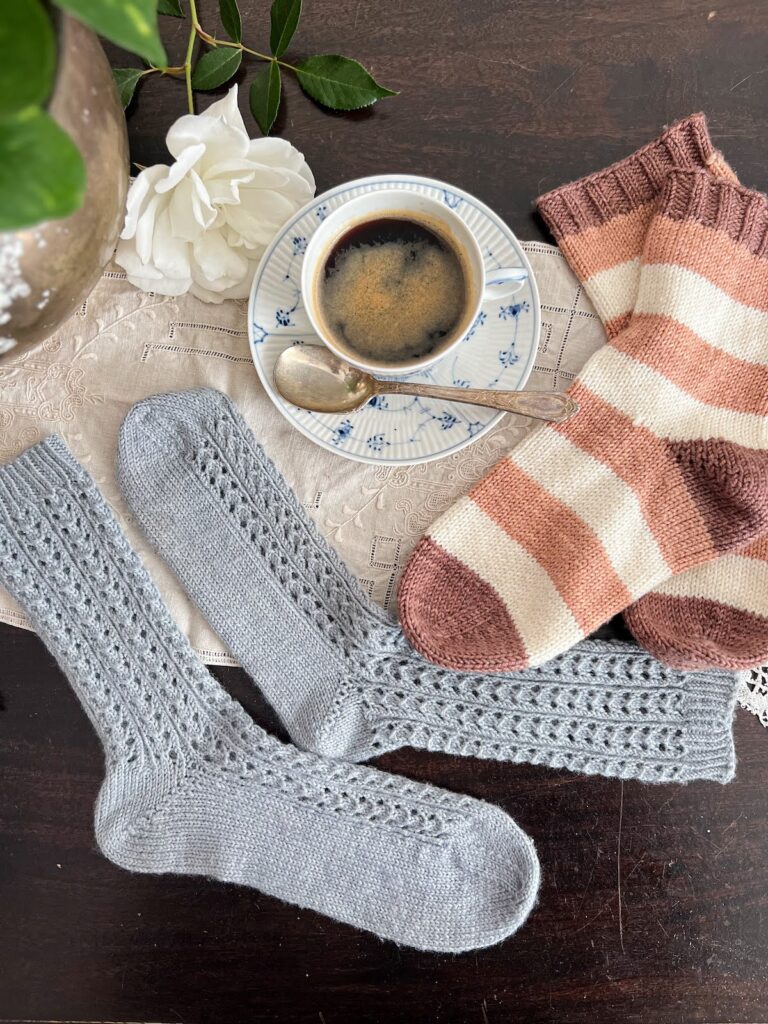 A top-down photo of the two pairs of socks featured in this post, one light blue and lacy, one with pink, white, and brown stripes. Both have had all their pills removed and look crisp and sharp again. Tucked in the middle between them is a blue and white teacup full of coffee and a white rose.