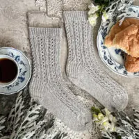 A flatlay photo of a pair of light gray handknit socks. The socks are laid flat on a gray surface with the toes pointing to the right. They're covered in tiny cables and eyelets, and surrounded by dusty miller, a blue and white teacup full of coffee, and a blue and white place with several croissants on it.