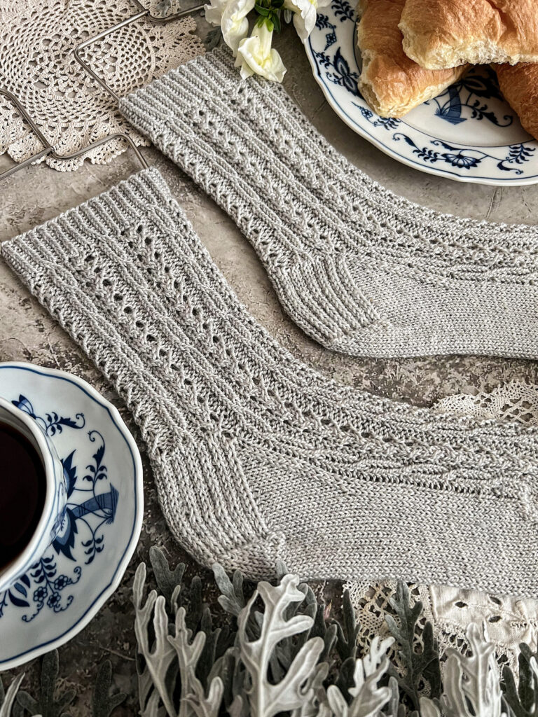 A detail shot of a pair of light gray handknit socks with lots of tiny cables and eyelets. The photo focuses on the slipped stitch heels and the delicate cables on the legs. You can see how the ribbing of the cuffs flows into the cables.