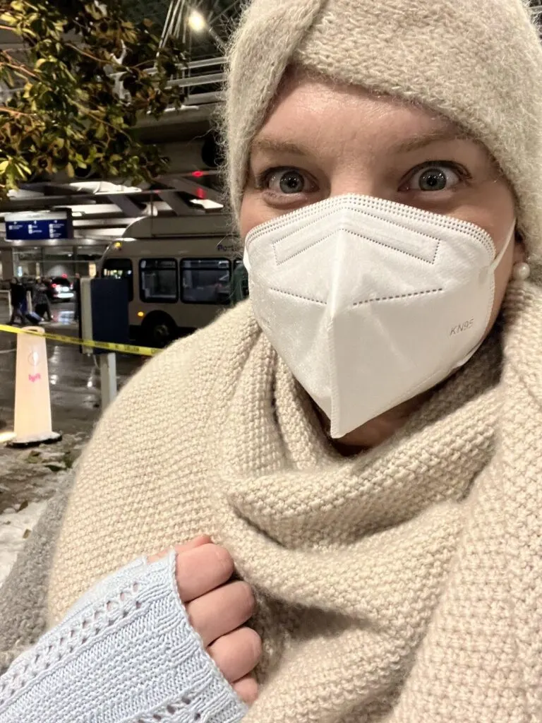 A freezing cold woman (yours truly) is bundled up in an earwarmer, knit shawl, and light blue fingerless mitts. I'm wearing a white KN95 mask and my eyes are open very wide because it is VERY COLD OUTSIDE