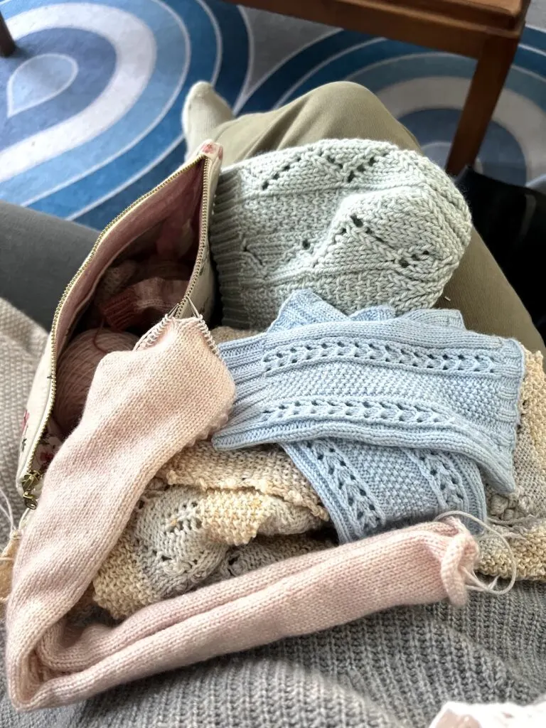 A pile of knits in my lap while I sit on the sofa. There's a pale pink earwarmer in progress, a light green knit hat, light blue fingerless mitts, and a pale yellow and light blue shawl.