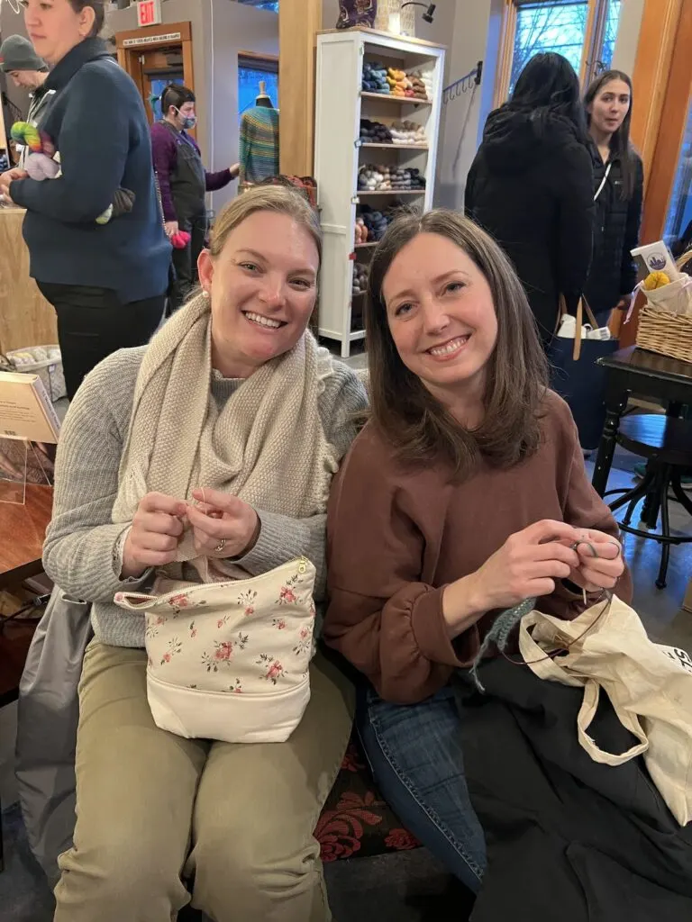 Two women sitting together on an oversized chair, knitting together in a yarn shop. One is me, and one is my friend Emily. Our heads are leaning together and we're both wearing very big smiles.