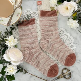 A flatlay photo of a pair of plain handknit socks. The socks are knit in a softly variegated pink and cream yarn, with contrasting cuff and toes in darker pink. The toes point to the right. The socks are surrounded by greenery, white flowers, and a white teacup filled with milky Earl Grey.