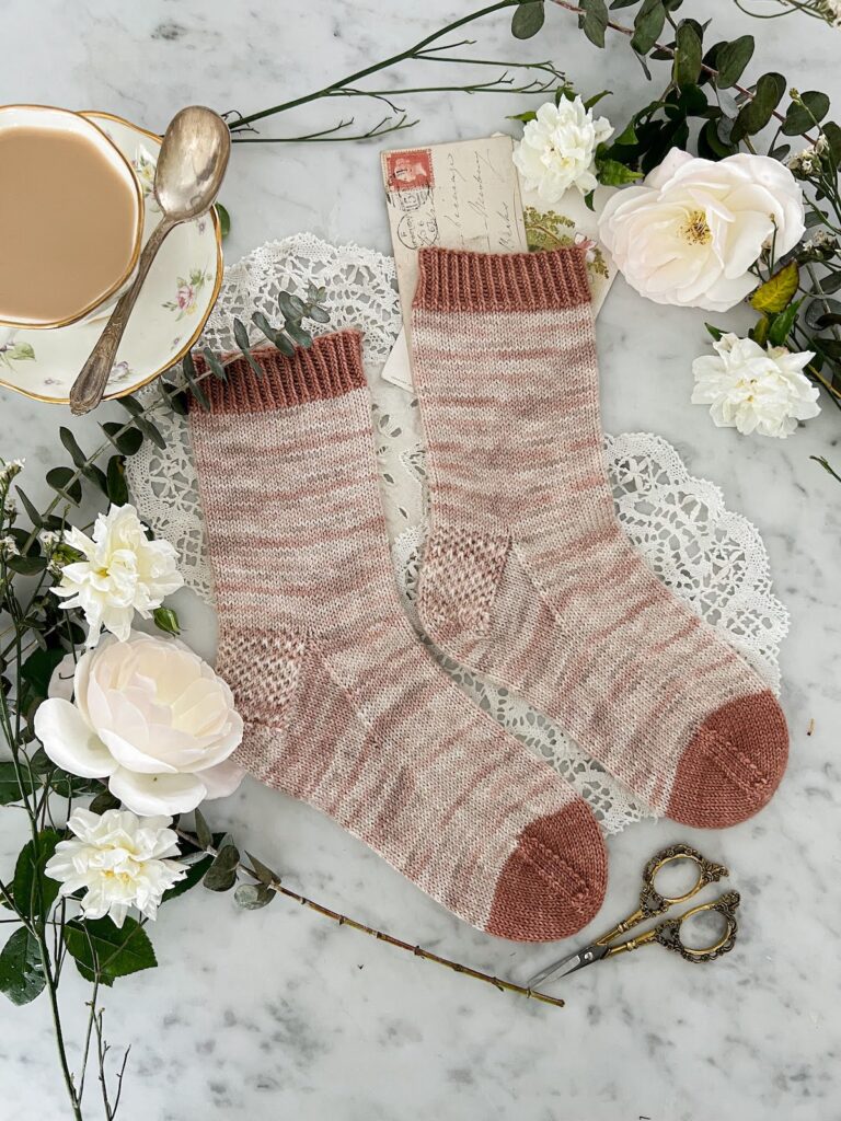 A flatlay photo of a pair of plain handknit socks. The socks are knit in a softly variegated pink and cream yarn, with contrasting cuff and toes in darker pink. The toes point to the right. The socks are surrounded by greenery, white flowers, and a white teacup filled with milky Earl Grey.