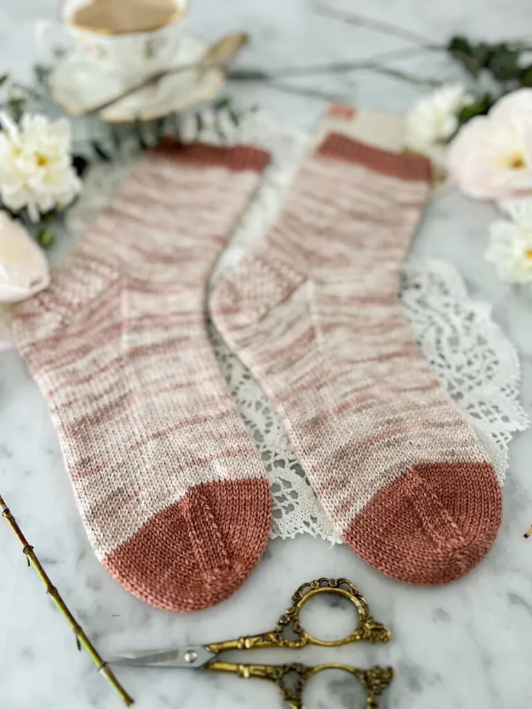A pair of plain, handknit socks in a softly variegated pink and cream yarn is laid flat on a countertop. The image zooms in on the toes, which are knit in a contrasting deep pink yarn.