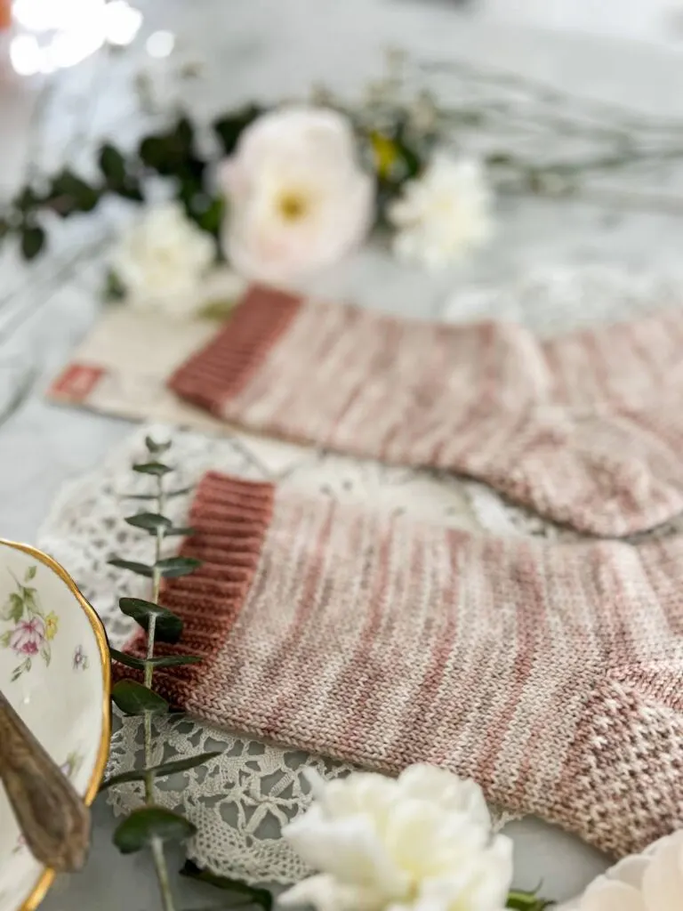 A close-up on the cuff of the left sock. It is knit in a contrasting pink color in a 1x1 twisted rib. The leg of the sock is knit in stockinette stitch using a softly variegated pink and cream yarn.