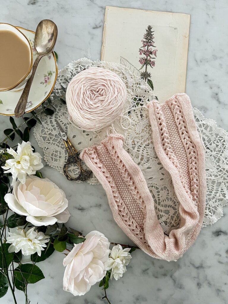 A top-down photo of a tube of light pink knitting with columns of eyelets and seed stitch surrounded by white roses, antique doilies, a vintage botanical print, and a white teacup full of milky tea.