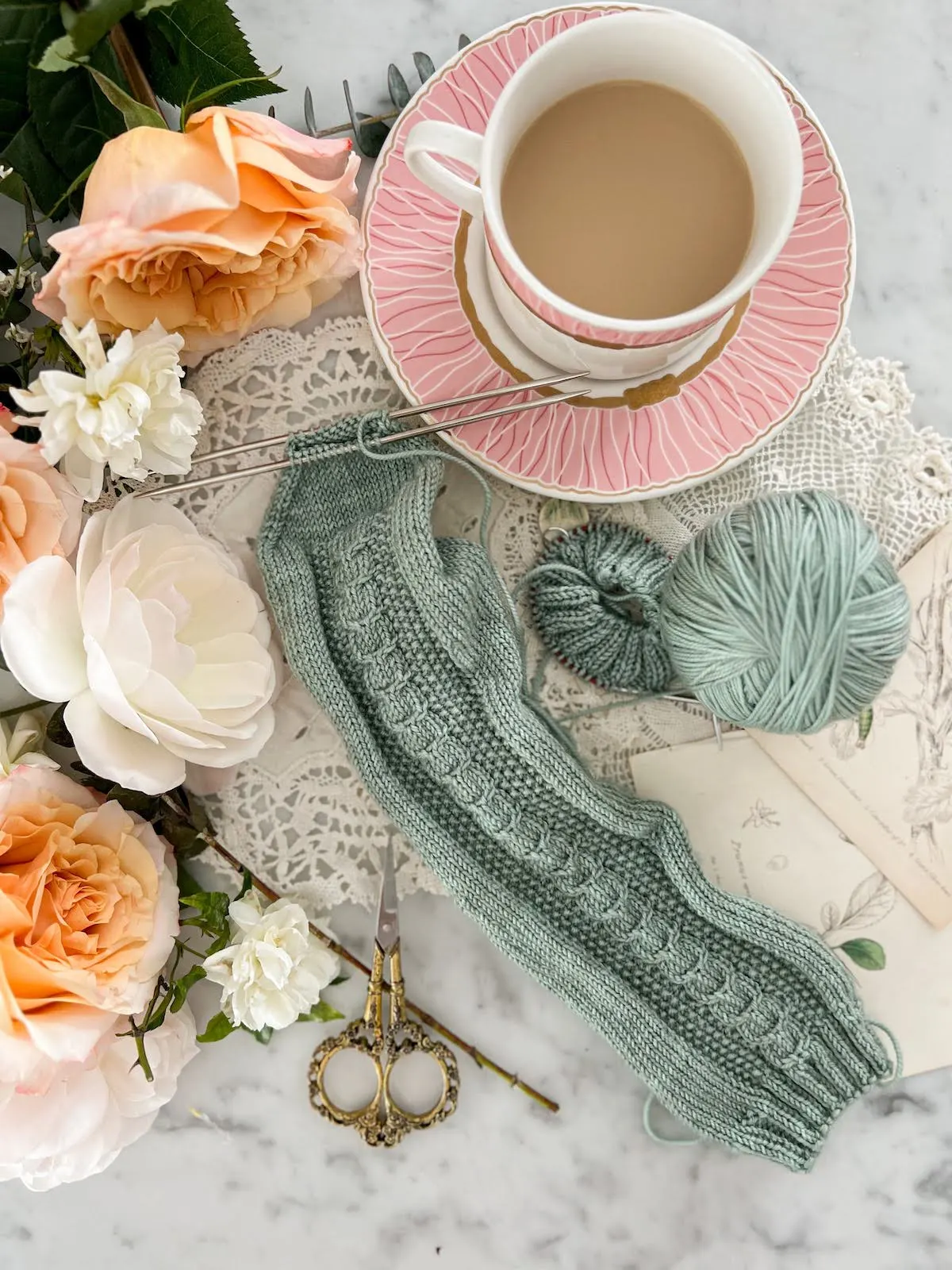 A top-down photo focused on a pair of handknit sock in progress. One sock is almost done and just needs the toes grafted, while the other is just ribbing on a small circular needle. The socks are a soft blue-green with hints of gray and feature a cabled panel around the front. They're surrounded by a pink teacup and saucer (with lots of milky tea in the cup), antique doilies, and peach and white roses.