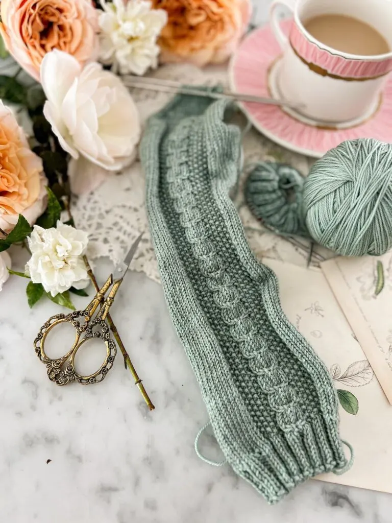 A close-up on the leg portion of a pale green-blue sock in progress. There is a small cabled panel down the front. In the background are peach and white roses and a pink teacup full of milky tea.