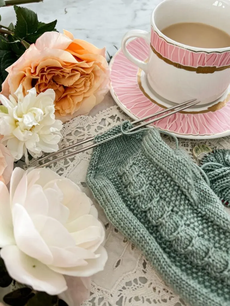 A close-up on the toe of an almost-finished sock. The toe stitches are arranged on two needles and ready to be grafted together. The sock is surrounded by a pink teacup full of milky tea and some peach and white roses.