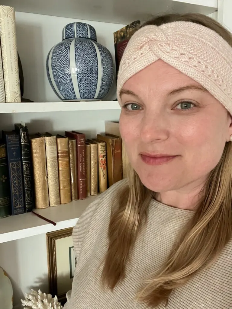 I'm wearing my finished headband and a tan top in front of a bookcase in my living room. The headband is a pale pink and wraps over my ears with a little twisty knot feature at the top of my head. I've got a soft smile in this photo because I'm very happy with the headband but still feel a bit silly taking pictures of myself.