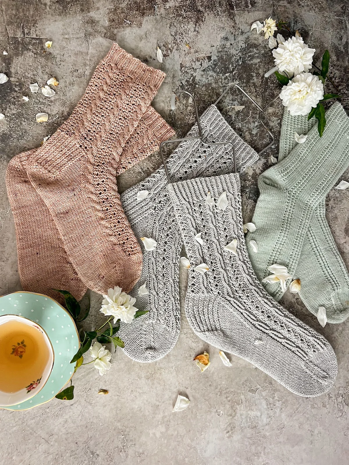 A flatlay photo showing a pair of pink cabled socks with eyelets on the left, a pair of blue-gray cabled socks in the middle, and a pair of mint green socks with columns of eyelets on the right. The blue-gray socks are on a pair of wire sock blockers. A mint green teacup full of chamomile and a few small white roses complete the scene.