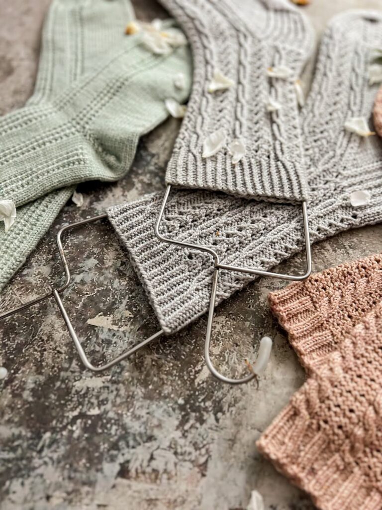 A slightly jumbled pile of pastel socks, with slight snippers of pink and mint green socks peeking in on either side and a pair of blue-gray cabled socks taking up the majority of the photo. The image is focused sharply on the tops of the wire sock blockers used for the cabled socks.