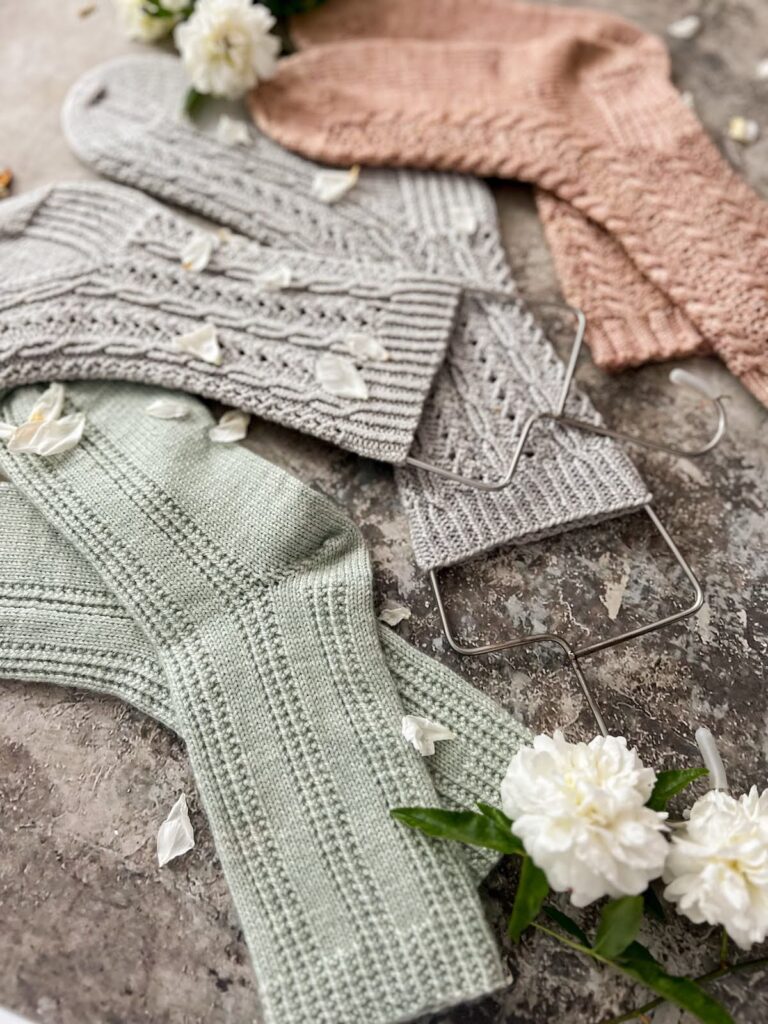 A pair of three pastel socks is laid out on a gray background. In focus in the foreground is a pair of mint green socks with columns of purl stitches. Slightly blurred in the middle of the photo is a pair of cabled blue-gray socks on wire sock blockers. In the background is a pair of pink cabled socks.