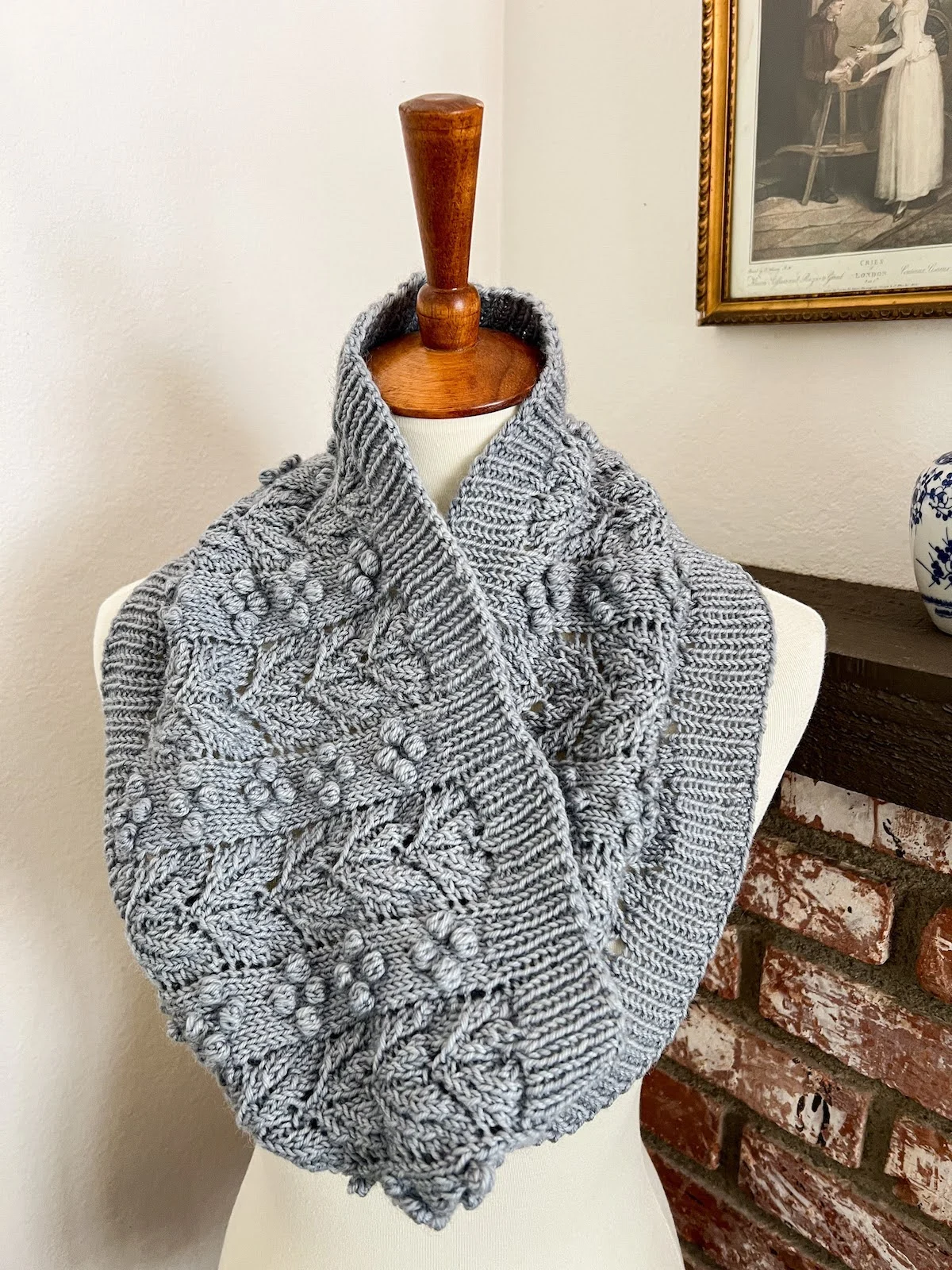 A light blue wool cowl is draped around the neck and shoulders of a dressmaker's form. The cowl features alternating columns of bobbles and lacy leaves, with 1x1 ribbing along the top and bottom for stability.