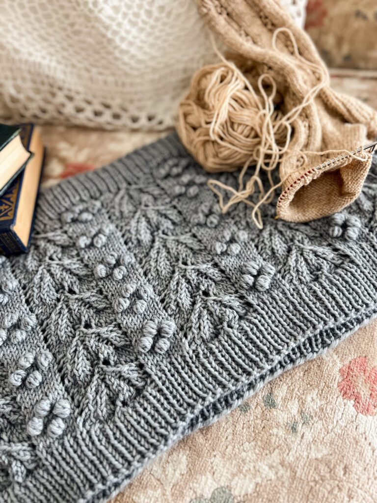 A light blue wool cowl is spread across the seat of a tan sofa with with a soft floral print. In the background are some antique books and a knit sock in progress.