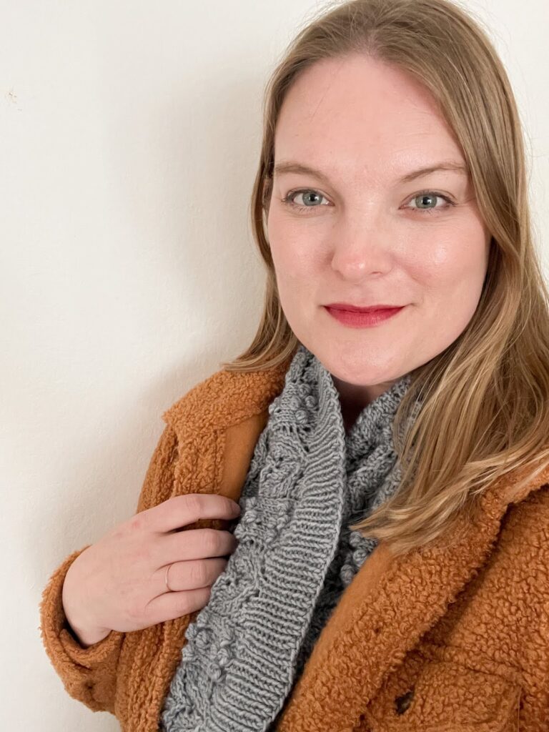 A blonde woman in her late 30s (me!) smiles softly at the camera. She's wearing a butterscotch-colored fleece jacket and a blue knit cowl.