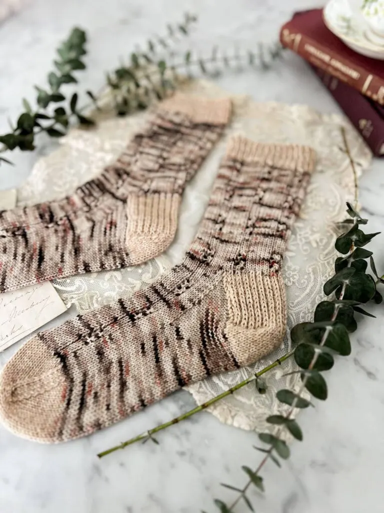 A pair of brown, pink, and gray speckled socks with contrasting tan cuff, heels, and toes are laid out flat on a countertop. The image is shot low from the heel of the right sock across toward the left sock, which blurs into the background. This image shows the construction of the whole foot with special attention on the slipped stitch flap heel.