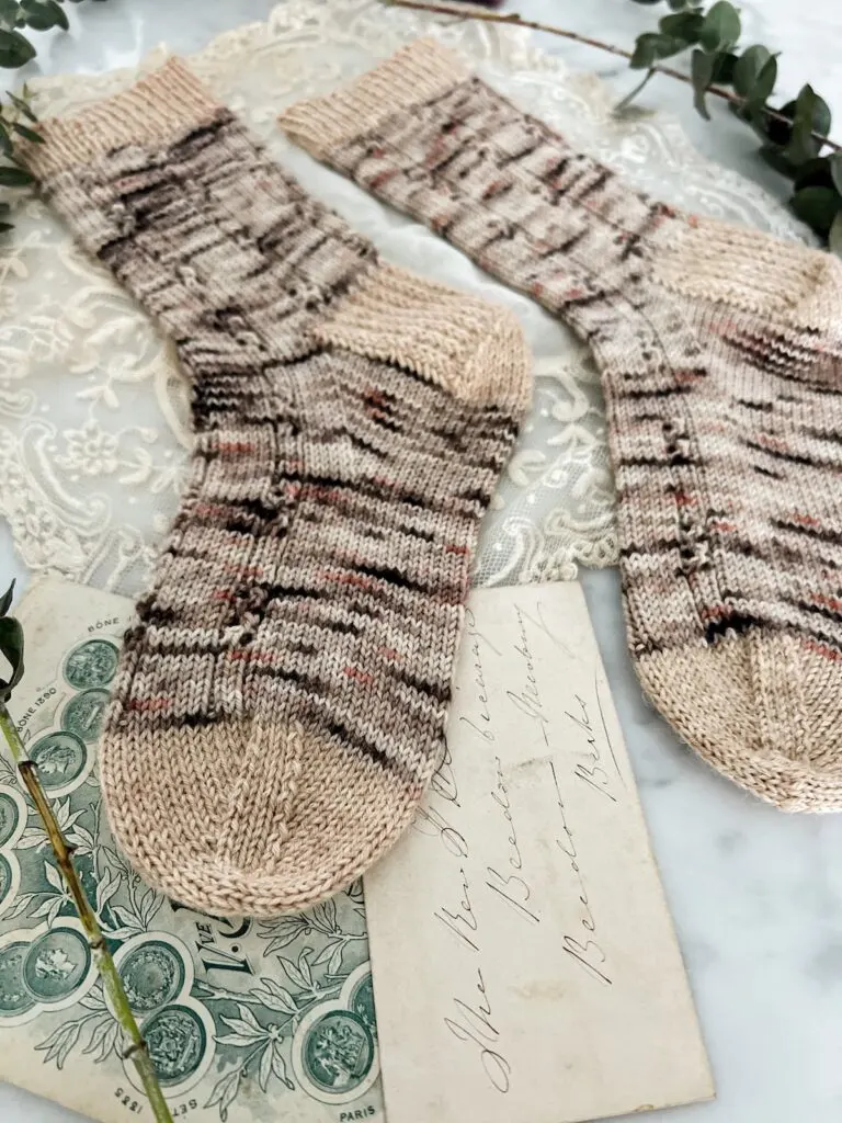 A pair of brown, pink, and gray speckled socks with contrasting tan cuff, heels, and toes are laid out flat on a countertop. The image is shot low from the toe of the left sock up along the feet, which blur into the background. This image gives a detailed look at the wedge toe construction.