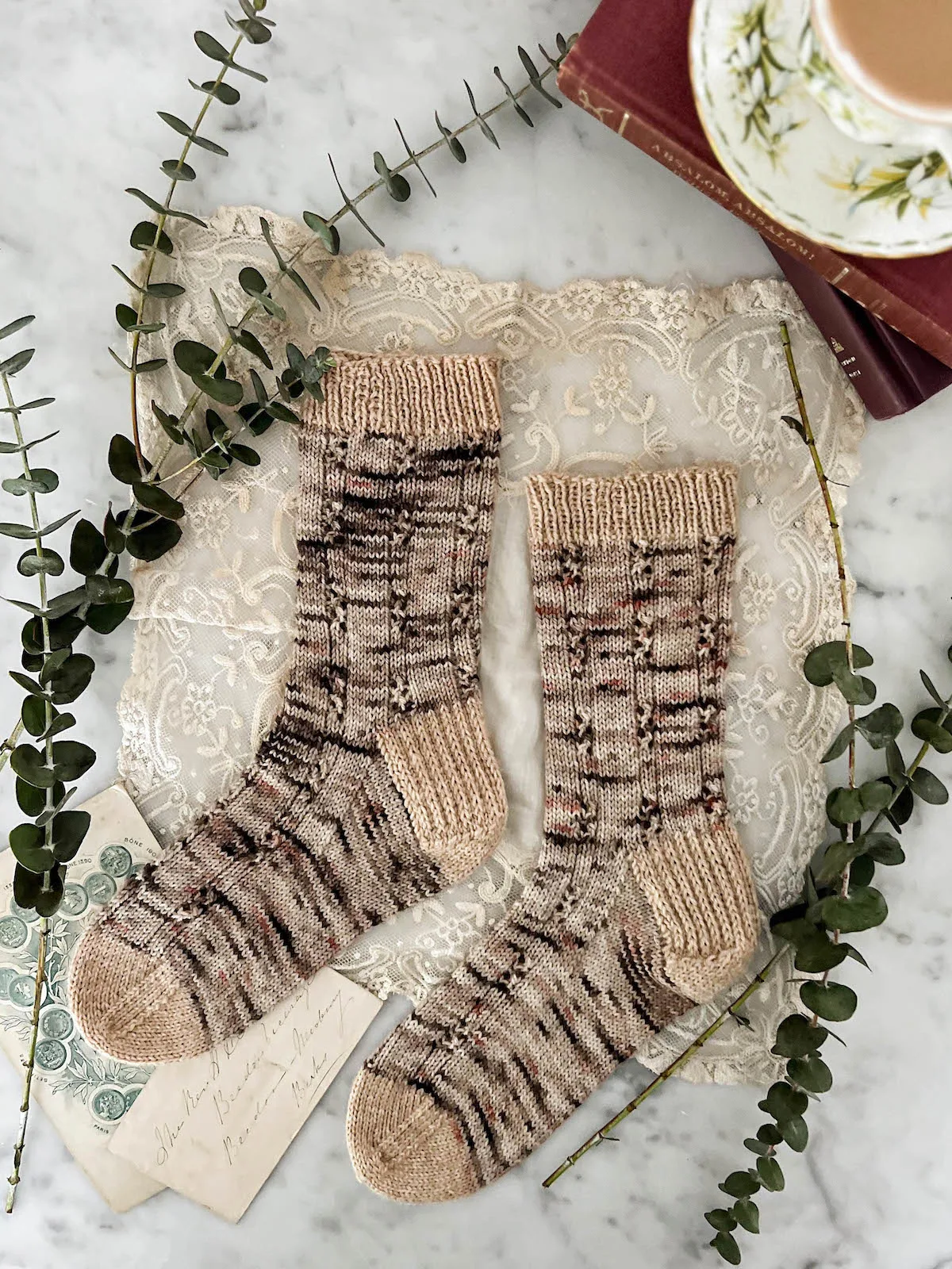 A pair of speckled socks is laid out out flat on a white marble countertop with the toes pointing to the left. The socks are knit in a pattern that alternates columns of stockinette stitch with columns of ribbed eyelets. The socks are surrounded by small eucalyptus branches, antique books, and a lace handkerchief.