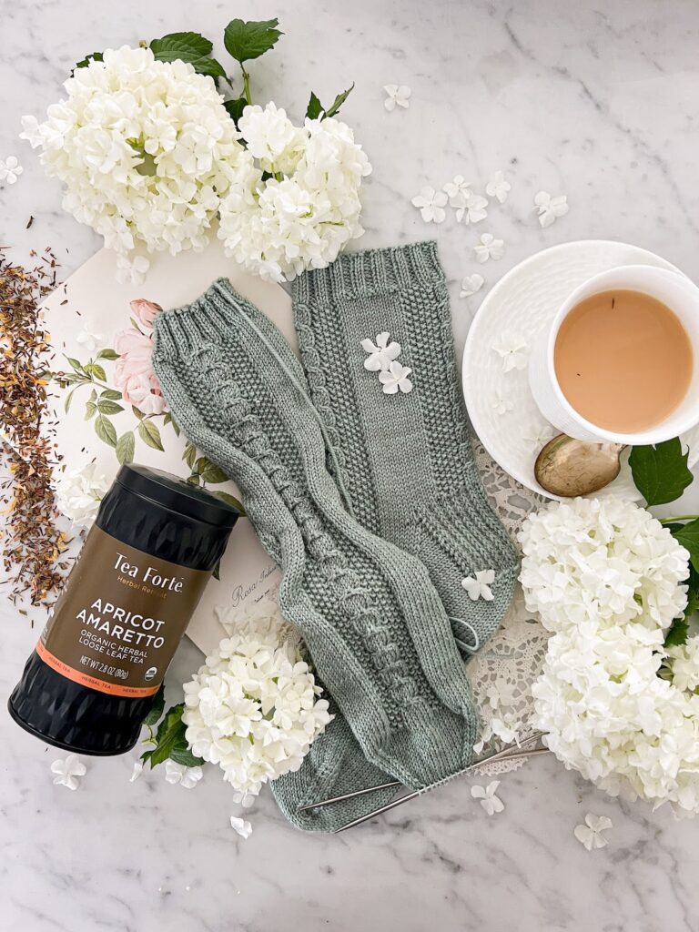 A top-down photograph of a pair of gray-green socks with cable and seed stitch panels down the front and back. One sock still needs its toe grafted together. To the left of the socks is a canister of Tea Forte's Apricot Amaretto tea. It's a black canister with a tan label that has apricot-colored embellishments. A little tea is spilled across the counter to show its texture. To the right of the socks is a white teacup and saucer. The cup is full of Apricot Amaretto tea with milk. Small balls of snowball viburnum are scattered about the image.