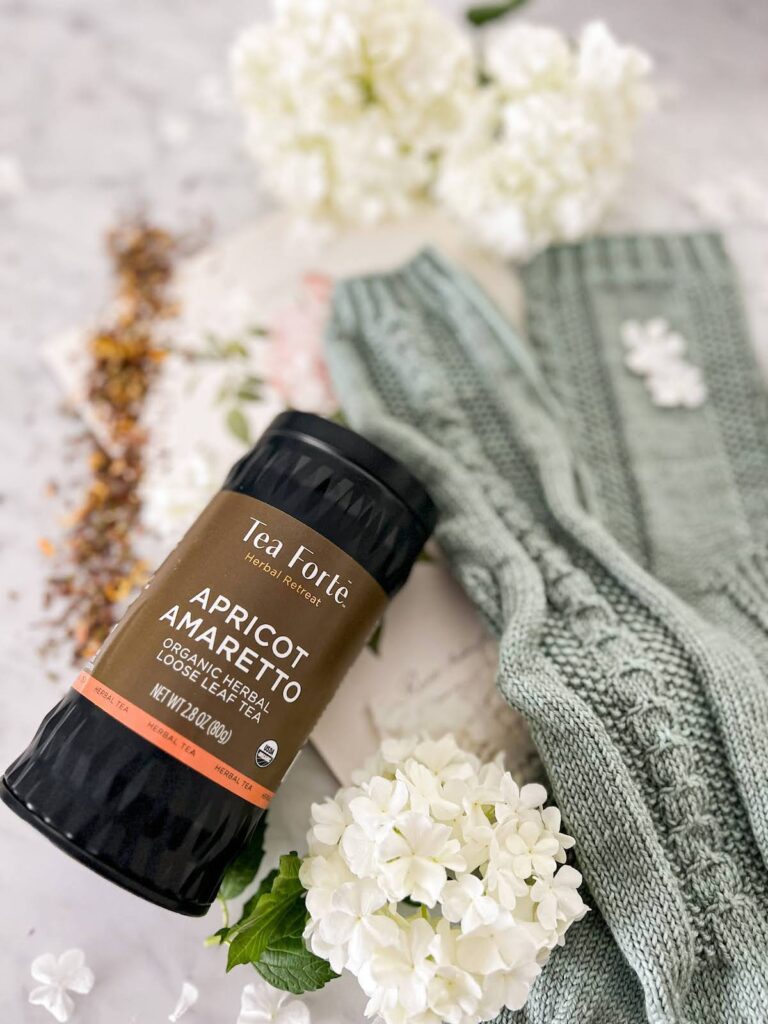 A close-up photo of a canister of Tea Forte's Apricot Amaretto tea. It is a black canister with a brown label that has apricot-colored accents. The canister is on its side with the label facing upward. Blurred in the background are some white flowers and some green-gray socks.