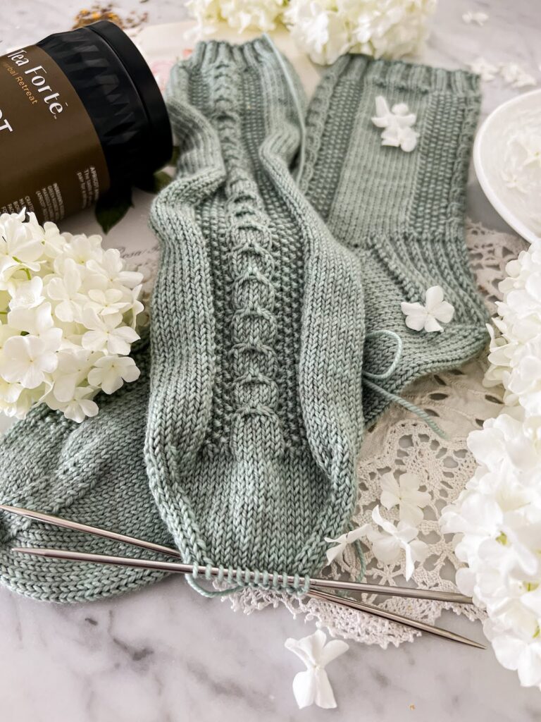 A close-up photo of the toe of a a gray-green sock with cable details and seed stitch going down the front of the sock. The toe is still on the needles and needs to be grafted shut.