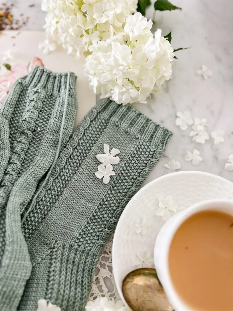 A close-up on the legs of a pair of gray-green socks with cable details and seed stitch panels down the front and back. To the top are some fluffy white flowers. In the bottom right is a white teacup full of milky tea.
