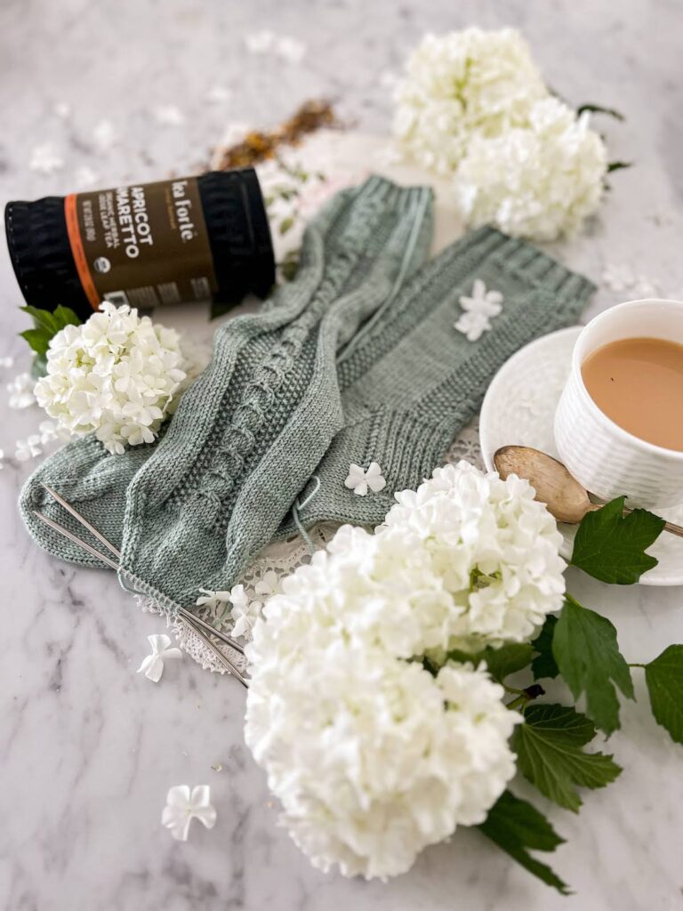 An angled image of a pair of gray-green socks with cable details and seed stitch panels down the front and back. This image has the toes in focus in the middle of the picture and blurs the leg of the sock as they move into the background. Blurred in the foreground are some puffy white flowers. In the background are a canister of tea and a white teacup full of milky tea.