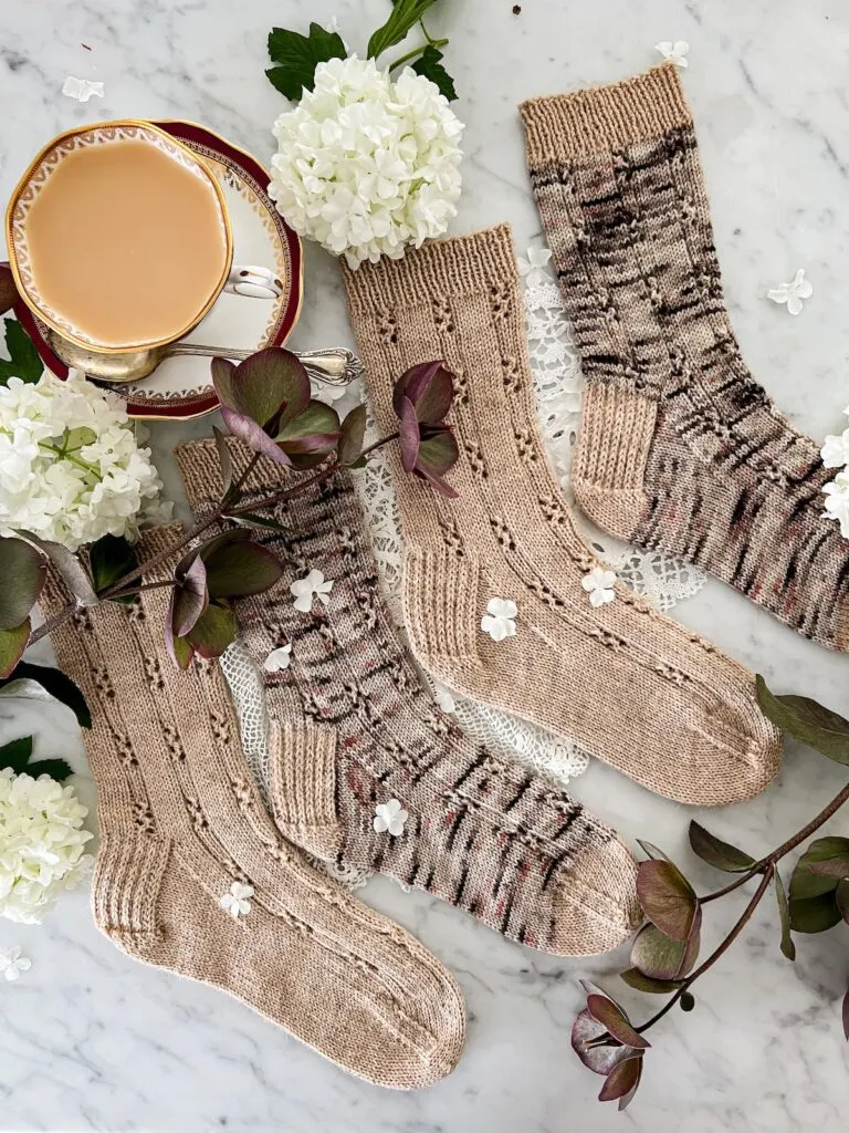 Two pairs of socks are laid out in an alternating pattern on a white marble countertop with the toes pointing to the right. One pair is knit in a tan yarn, while the other is knit in a brown, pink, and gray speckled yarn. The second pair has a contrasting cuff, heel, and toe knit with the same yarn as the first pair of socks. They are surrounded by a burgundy and white teacup full of milky tea, faded hellebores in shades of dark burgundy and green, and white puffy balls of snowball viburnum.