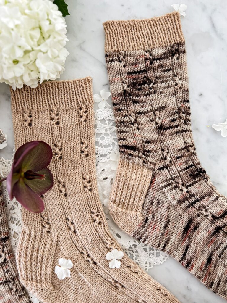 A top-down image of the legs of two socks knit in the same pattern of columns of stockinette broken up by ribbed eyelets. The sock on the left is all tan. The sock on the right is brown, gray, and pink speckled, with a contrasting cuff and heel knit in the same yarn as the first sock.