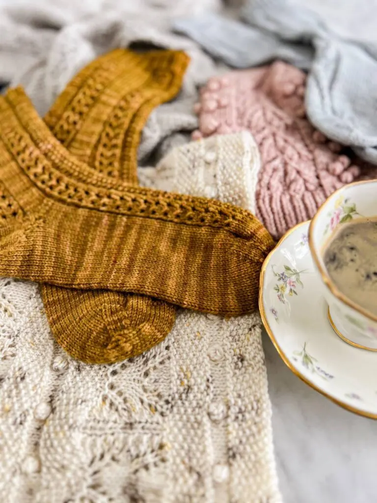A pair of caramel-colored handknit socks is in focus in the middle left of the photo. The socks are surrounded by other handknit accessories.