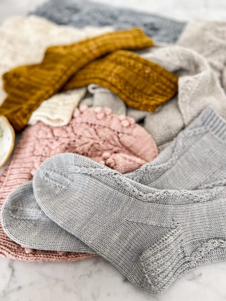 A pair of light blue handknit socks fill the foreground of this photo, which focuses on the front sock's heel and toe. Fading into the background are a pink hat, caramel-colored socks, a cream cowl with speckles, a gray shawl, and a blue cowl.