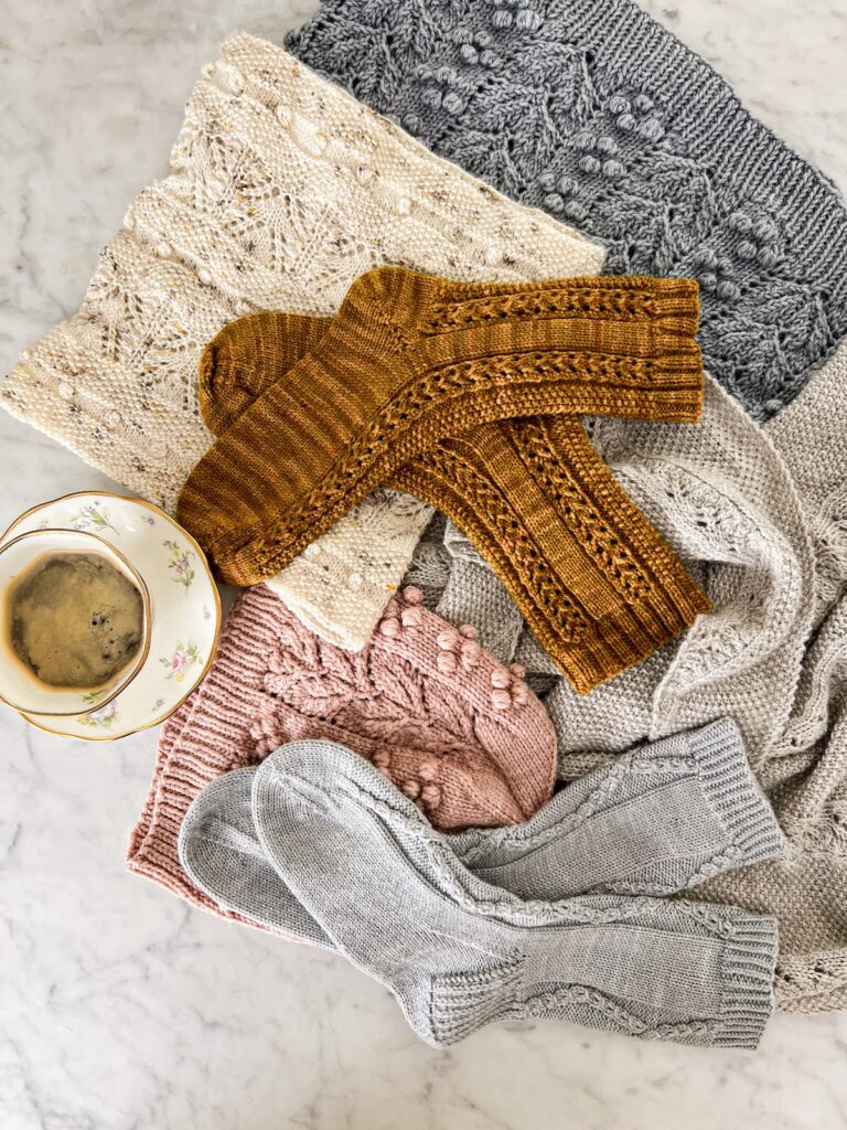 A pile of handknit accessories laid out on a white marble countertop with a white teacup full of espresso.